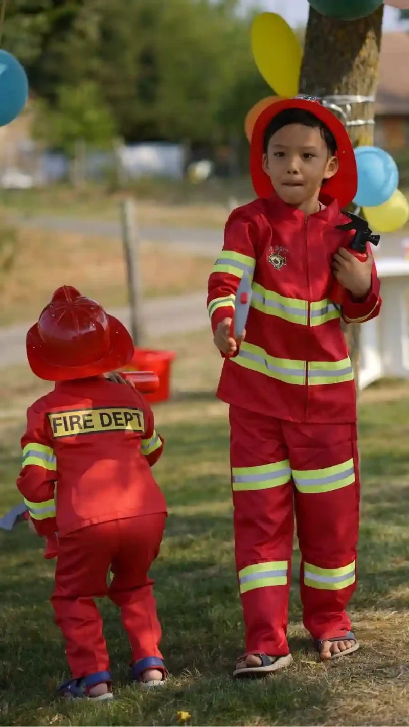 Deux enfants jouant aux pompiers dans le jardin, portant des costumes rouges réalistes avec l'inscription "FIRE DEPT" et des bandes réfléchissantes jaunes. Ils sont équipés de casques rouges et d'accessoires, prêts pour l'intervention.