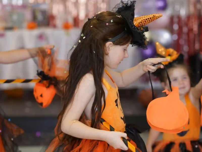 Petite fille aux cheveux longs lors d'une fête d'Halloween, portant une robe de sorcière orange et noire à bretelles confortables, un serre-tête chapeau pointu et tenant un sac citrouille pour les bonbons.