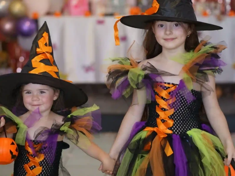 Deux petites filles se tenant la main, déguisées en sorcières avec des robes colorées en tulle (noir, orange, violet, vert) et des chapeaux pointus assortis, prêtes pour la fête d'Halloween.