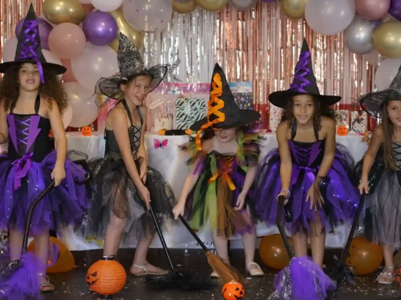 Groupe de cinq petites filles déguisées en sorcières lors d'une fête décorée de ballons. Elles portent des robes colorées en tulle (violet, noir, orange), des chapeaux pointus et tiennent joyeusement leurs balais magiques.