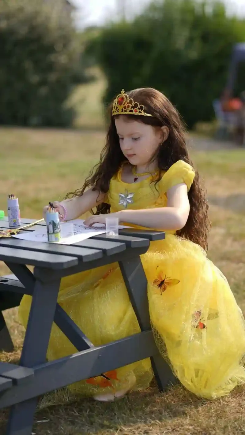 Petite fille aux longs cheveux bruns assise à une table de jardin, portant une robe de princesse jaune confortable ornée de papillons et une couronne, concentrée sur son coloriage.