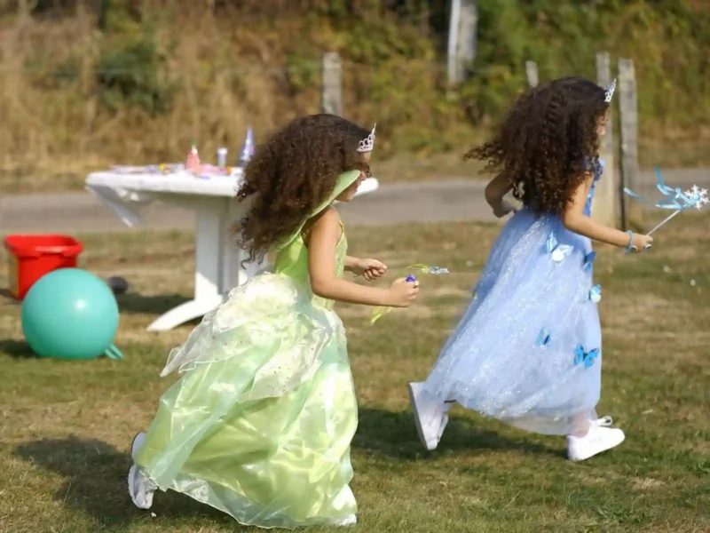 Deux petites filles courant joyeusement dans l'herbe lors d'un goûter d'anniversaire, portant des robes de princesse (verte et bleue) amples et résistantes, accessoirisées avec des baguettes magiques et des baskets.